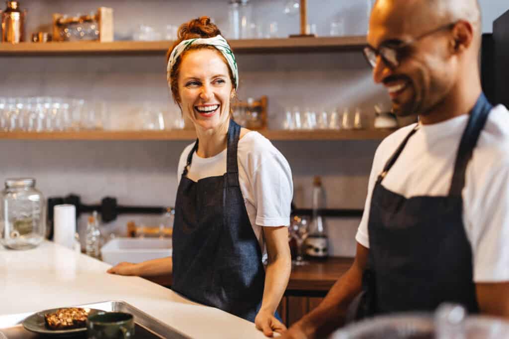 Two smiling baristas stand side by side behind the coffee bar, preparing to serve customers.