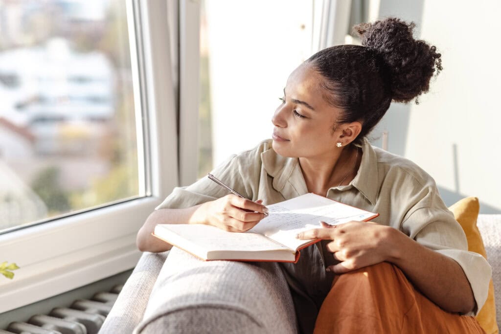 A young woman sitting on a sofa and smiling while looking out the window and thinking about what to write in her journal.