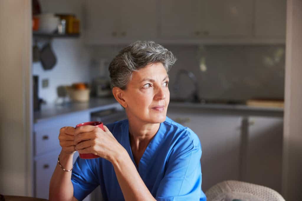 Woman with grey hair wearing scrubs sits in her kitchen holding a cup of coffee as the morning sunlight illuminates her face. She has a calm and grounded look on her face.