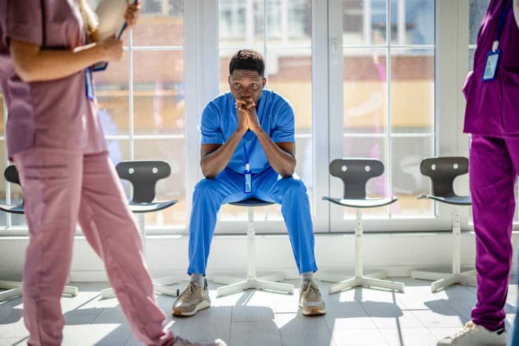 Male nurse siting in hall as other nurses walk by. They are deep on thought.