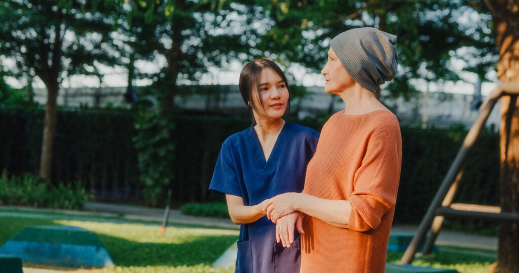 Cancer patient with headscarf walking with caregiver in park.