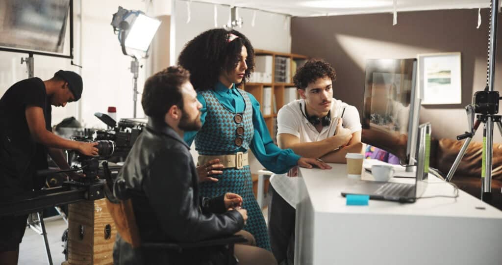 An actress and film production crew are standing on set and looking at a video monitor.