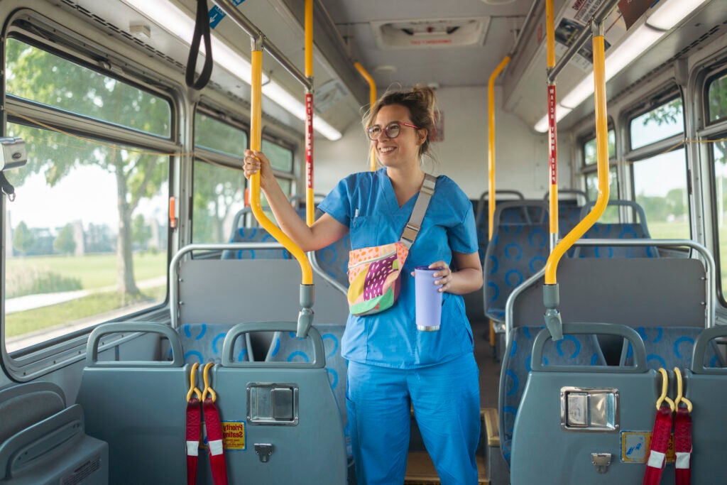 Smiling female nurse wearing blue uniform standing on empty bus holding travel cup going to work