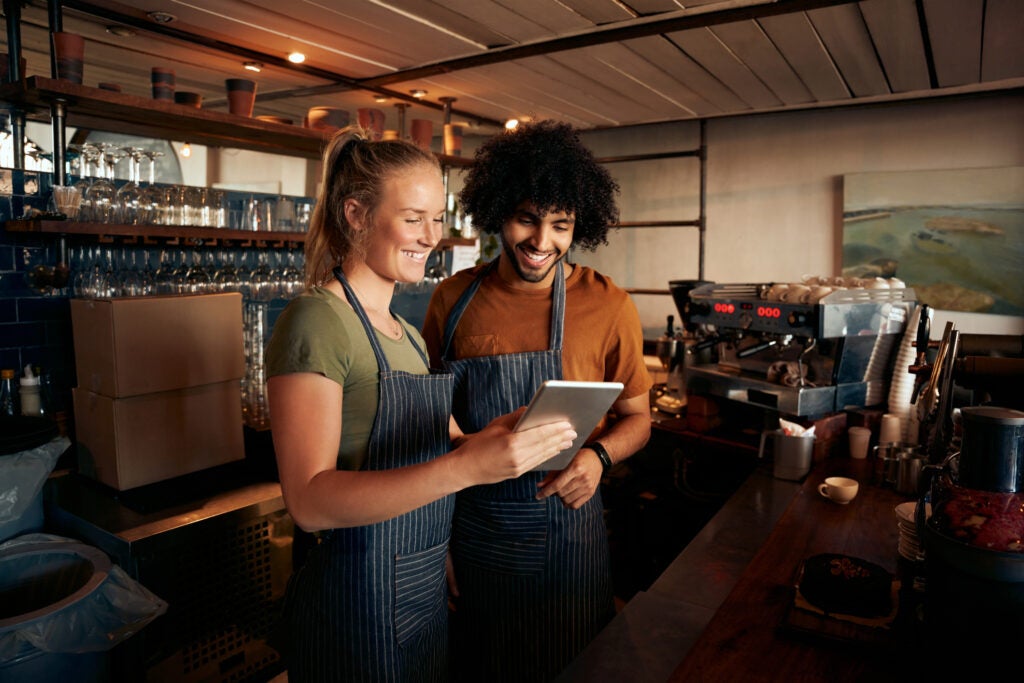 Smiling waiter and waitress wearing apron using digital tablet standing behind counter in cafe