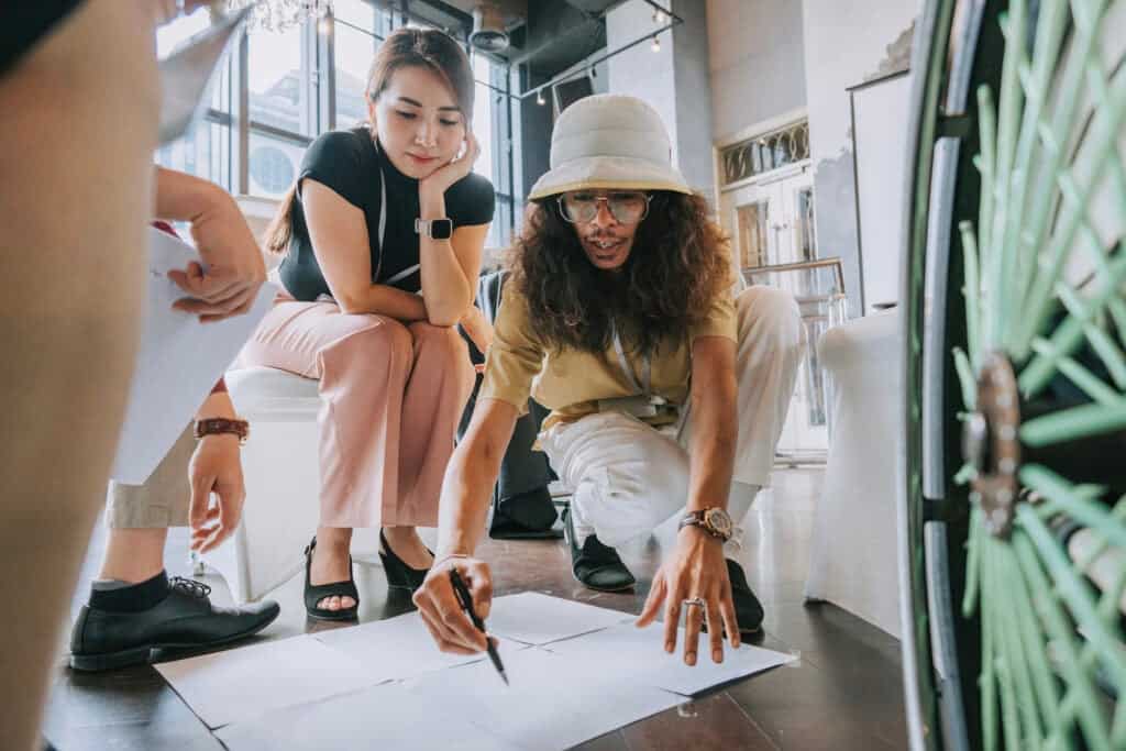 A diverse group of young creatives smiling and planning out a creative project by drawing plans on a large sheet of paper on the floor
